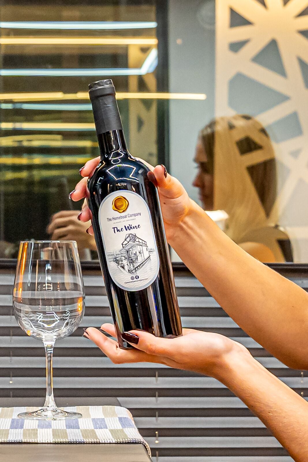 Una persona sostiene una botella de vino con una etiqueta que dice 'The Homeboat Company The Wine' junto a una copa de vino vacía sobre una mesa.