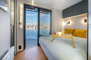 Una camera d'albergo moderna con un letto, un comodino in legno con articoli per la colazione, vista su una marina attraverso una porta in vetro e un piccolo balcone.