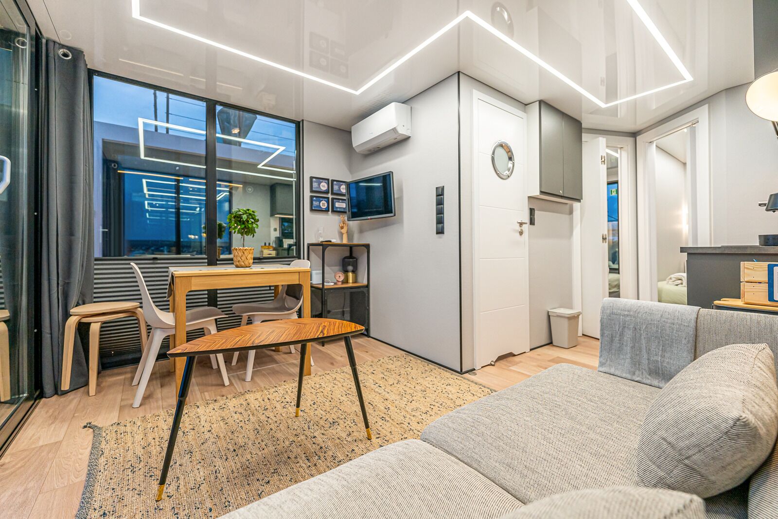 A modern, compact living space with a dining area, a sofa, a TV, and a small kitchen area.