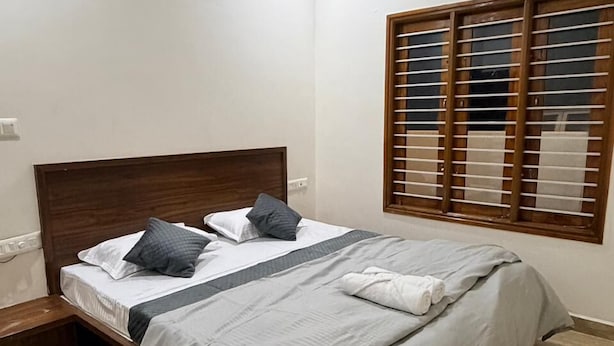 A neatly made double bed with a wooden headboard, a bedside table, and a window with wooden blinds. - The Residence 267 Homestay (Mysore)