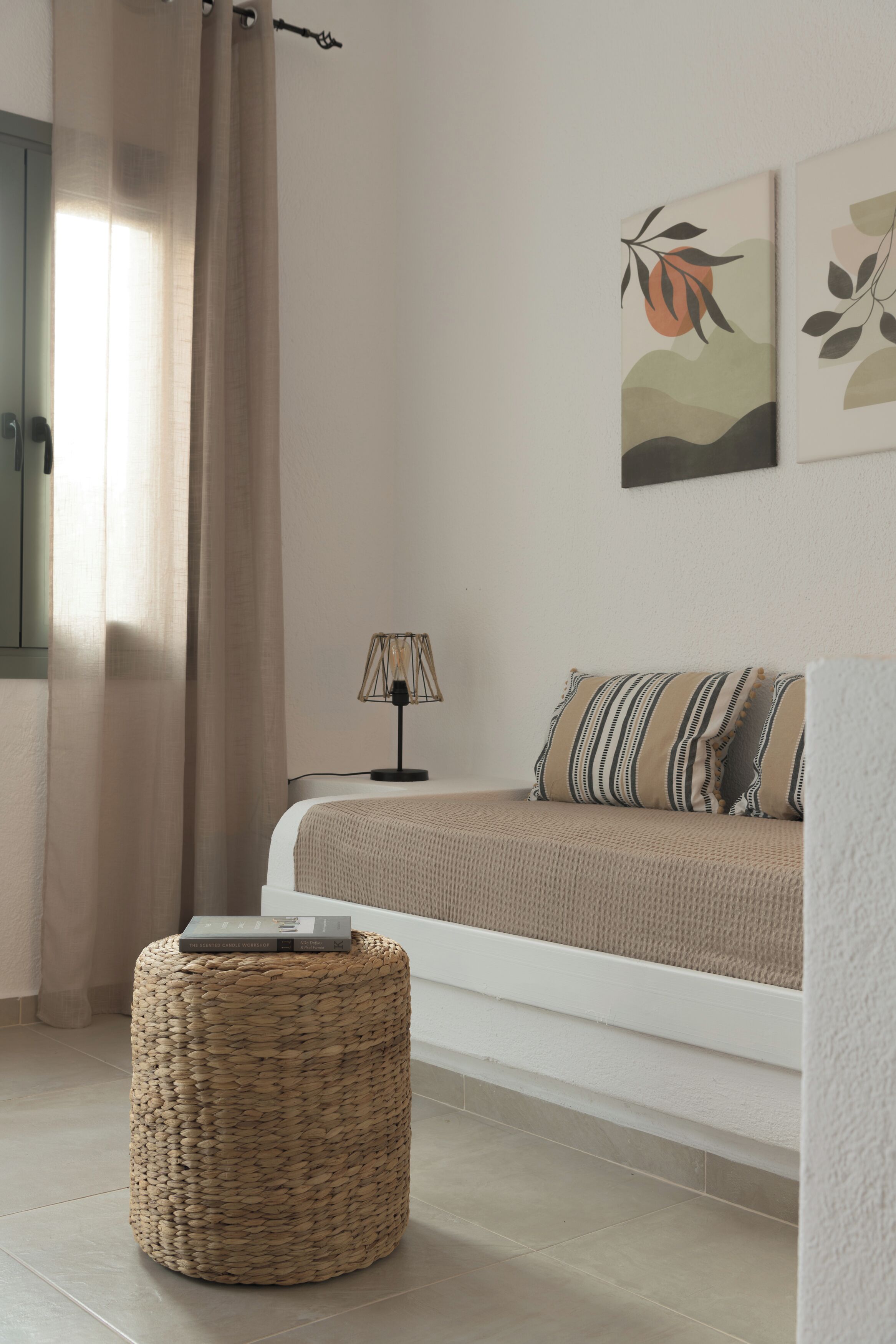 A bedroom with a bed, a bedside lamp, a woven ottoman, and wall art.