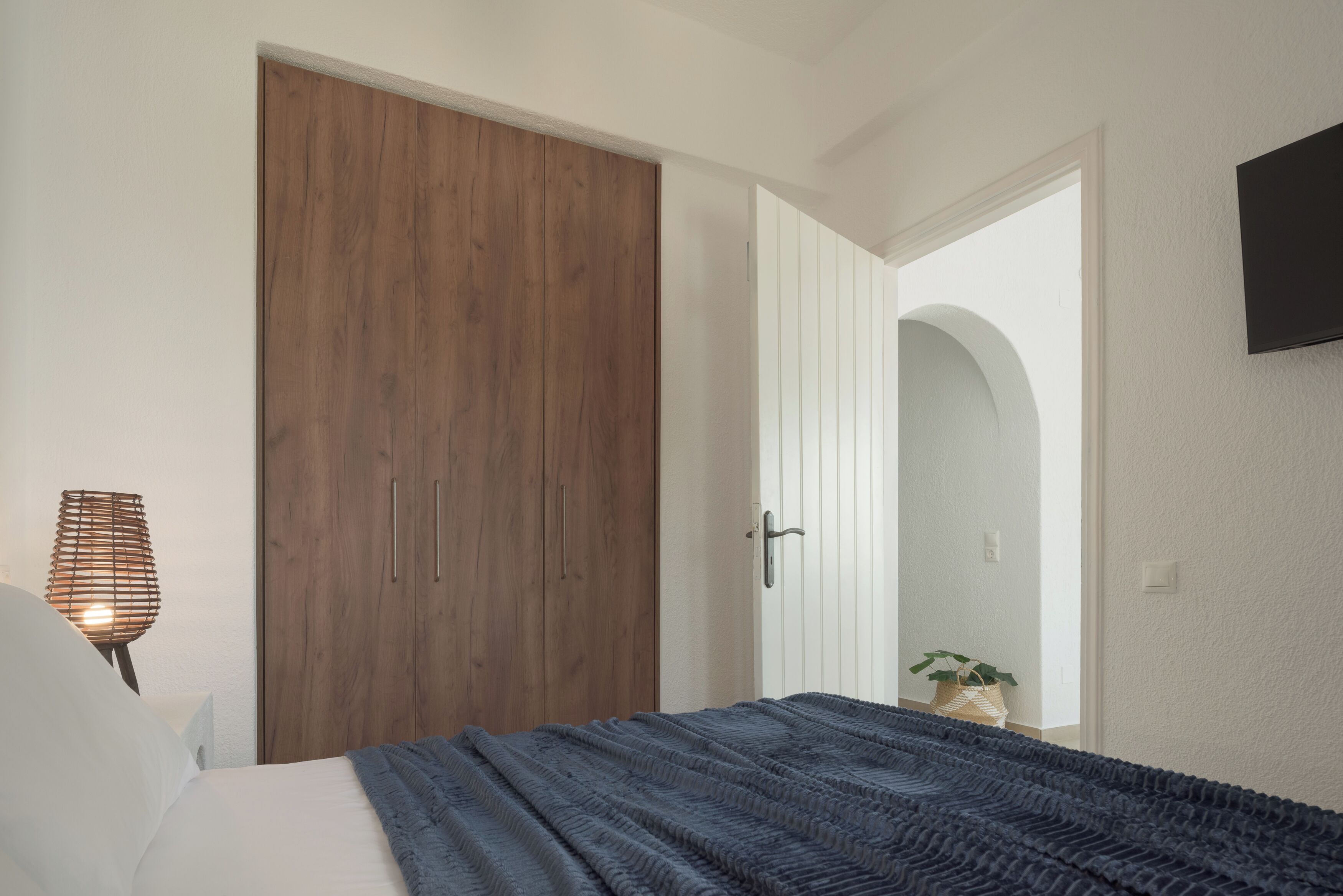 Un dormitorio con un armario de madera, una cama con manta azul, un televisor instalado en la pared y una lámpara de noche.