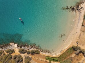 An aerial view of a coastal area with a boat on the water, a sandy beach, rocky outcrops, and some vegetation. - Voulisma Hills (Agios Nikolaos)