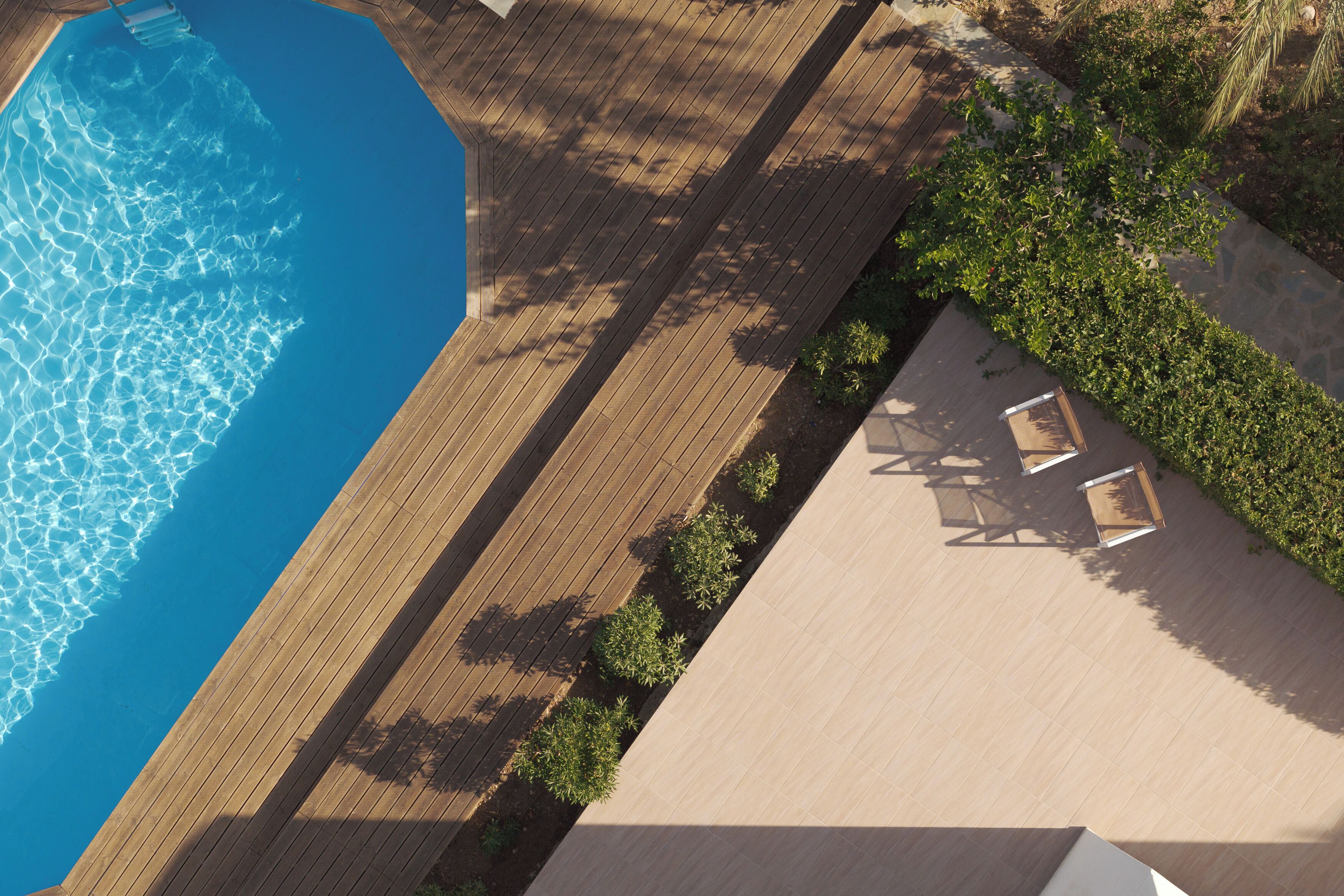 Vista aérea de un área de piscina con terraza de madera, zona de descanso con sillas y jardín ajardinado.