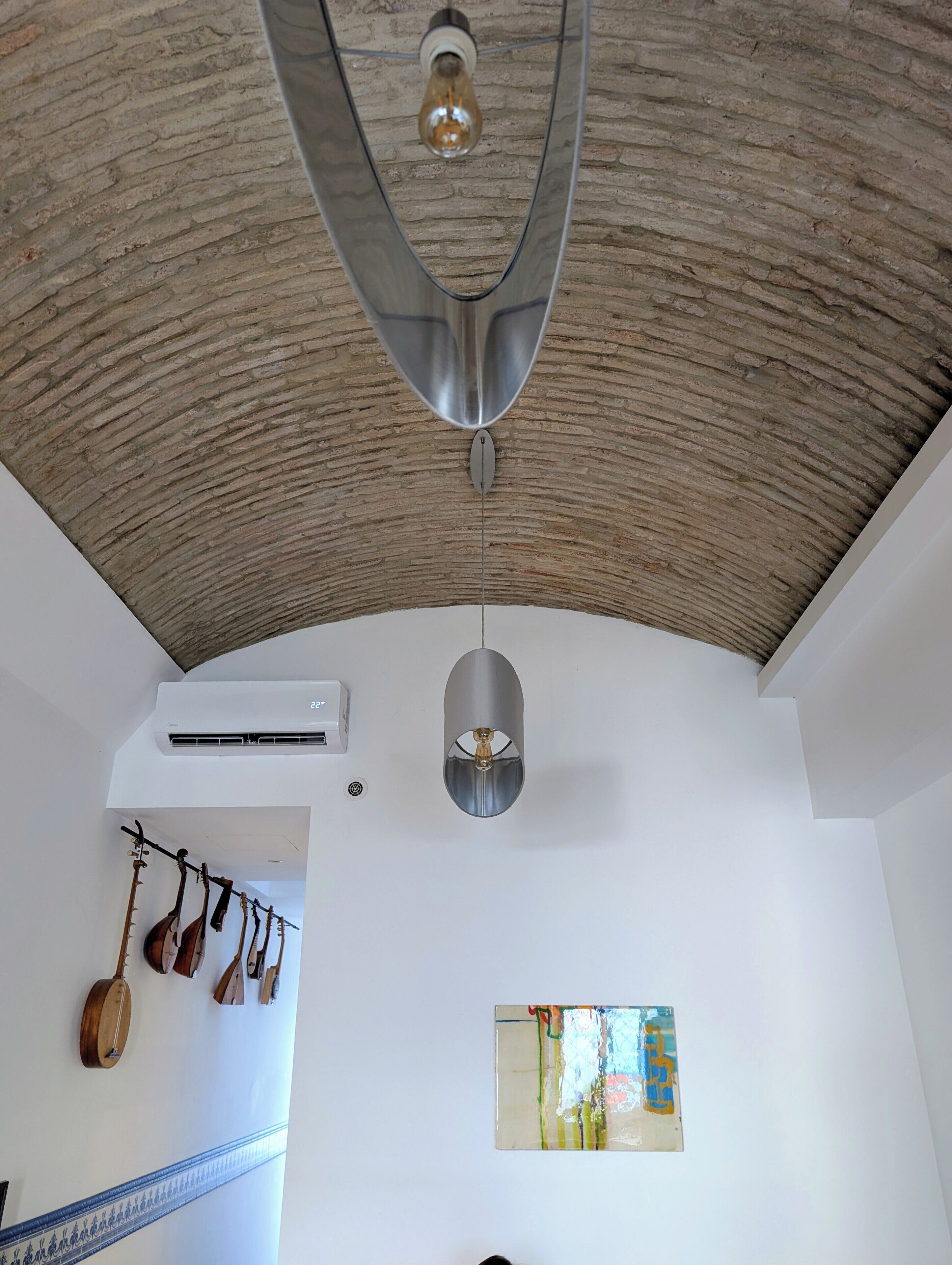 A room with a vaulted ceiling, a hanging light fixture, a wall-mounted air conditioner, and a string of musical instruments.