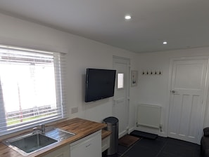 Eine Küche mit Holzarbeitsplatte, Edelstahlspüle und Geschirrspüler. Ein Fernseher ist an der Wand montiert, es gibt eine weiße Tür und ein Fenster mit Jalousien.