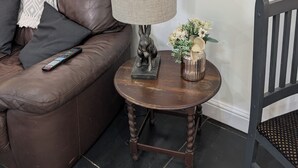 A brown leather sofa with a remote control, a round wooden side table with a lamp, a small statue, and a vase with flowers. - Quiet self contained apartment with free parking, close to Hospital (Torquay)