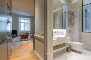 Ein modernes Badezimmer mit Waschbecken, Spiegel und Toilette in Weiß. Angrenzend befindet sich ein Wohnbereich mit einem Holzstuhl und einem Fernseher.