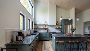 A modern kitchen with a large island, bar stools, and a view of the outdoors. - Modern Cabin | Hot Tub + Forest Views | 2 BR 2 BA (Logan)