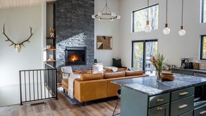 A modern kitchen with a central island, a stone fireplace, and a dining area with a wooden ceiling. - Modern Cabin | Hot Tub + Forest Views | 2 BR 2 BA (Logan)