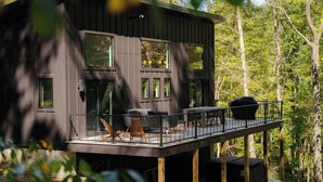 A modern cabin with a deck, large windows, and outdoor seating. - Modern Cabin | Hot Tub + Forest Views | 2 BR 2 BA (Logan)