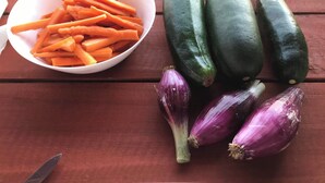 Puinen pöytä, jonka päällä on kulho viipaloituja porkkanoita, useita kokonaisia kurkkuja ja kaksi kokonaista punaista sipulia.
