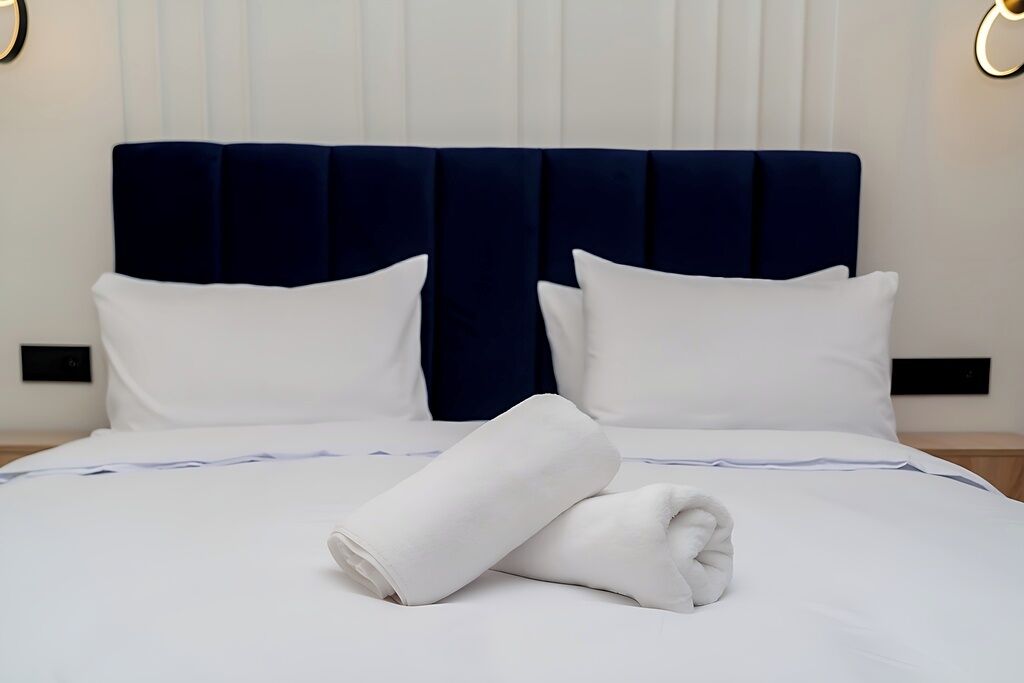 Hotellihuone, jossa on suuri sänky, valkoiset vuodevaatteet, tummansininen pääty ja sängyllä rullalle taitettu valkoinen pyyhe.