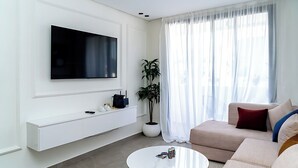 Ein modernes Wohnzimmer mit einem an der Wand montierten Flachbildfernseher, einem weißen Konsolentisch, einem runden Couchtisch, einem beige farbenen Sofa und einem großen Fenster mit transparenten Vorhängen.