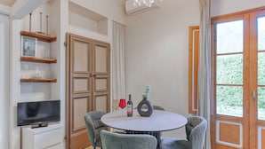 Uma sala de jantar com uma mesa redonda, quatro cadeiras, uma televisão sobre um móvel e uma janela com cortinas.