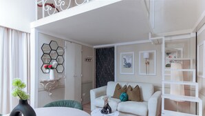 Uma sala de estar moderna com um sofá branco, poltronas verdes, uma mesa de centro redonda e uma mesa de jantar branca. Há um espelho hexagonal na parede, uma cortina preta e uma estante branca com objetos decorativos.