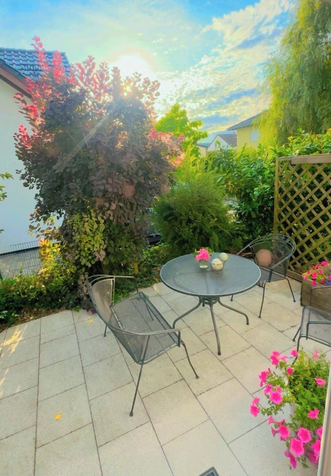 Eine Terrasse mit rundem Tisch und Stühlen, umgeben von Grünpflanzen und einem Gitterzaun.