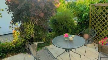 Eine Terrasse mit rundem Tisch und Stühlen, umgeben von Grünpflanzen und einem Gitterzaun.