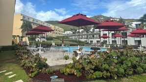 A pool area with red umbrellas, lounge chairs, and a view of hotel buildings and a mountain in the background. - Luxury Beachfront 3-Bedroom Condo | Ocean View (sint maarten)