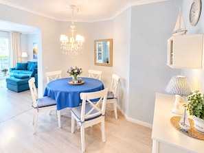 A dining area with a round table, white chairs, and a blue tablecloth. There is a chandelier above the table and a clock on the wall. - MeerZeit - cozy vacation apartment in Zingst - only 200 meters to the beach (Zingst)