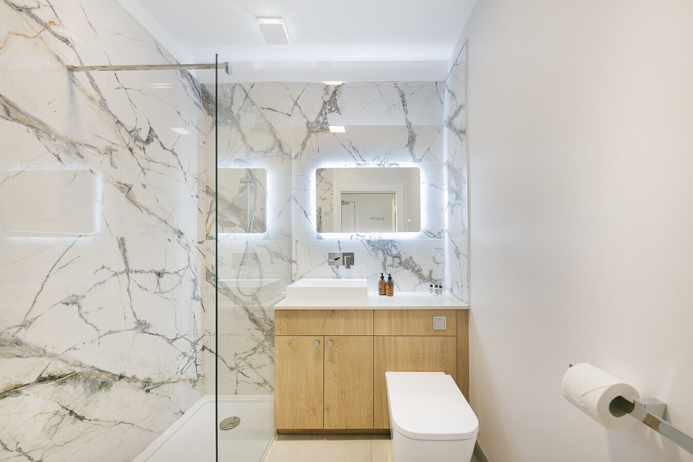A modern bathroom with marble walls, a glass shower enclosure, a wooden vanity, and a toilet.