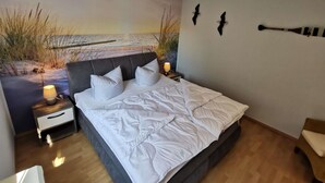 Une chambre avec un grand lit, deux tables de chevet avec des lampes, une fresque murale représentant une plage et un plancher en bois.