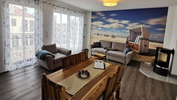 Ett vardagsrum med ett matbord i trä, två soffor, ett stort fönster med blommiga gardiner och en väggmålning med ett strandmotiv.