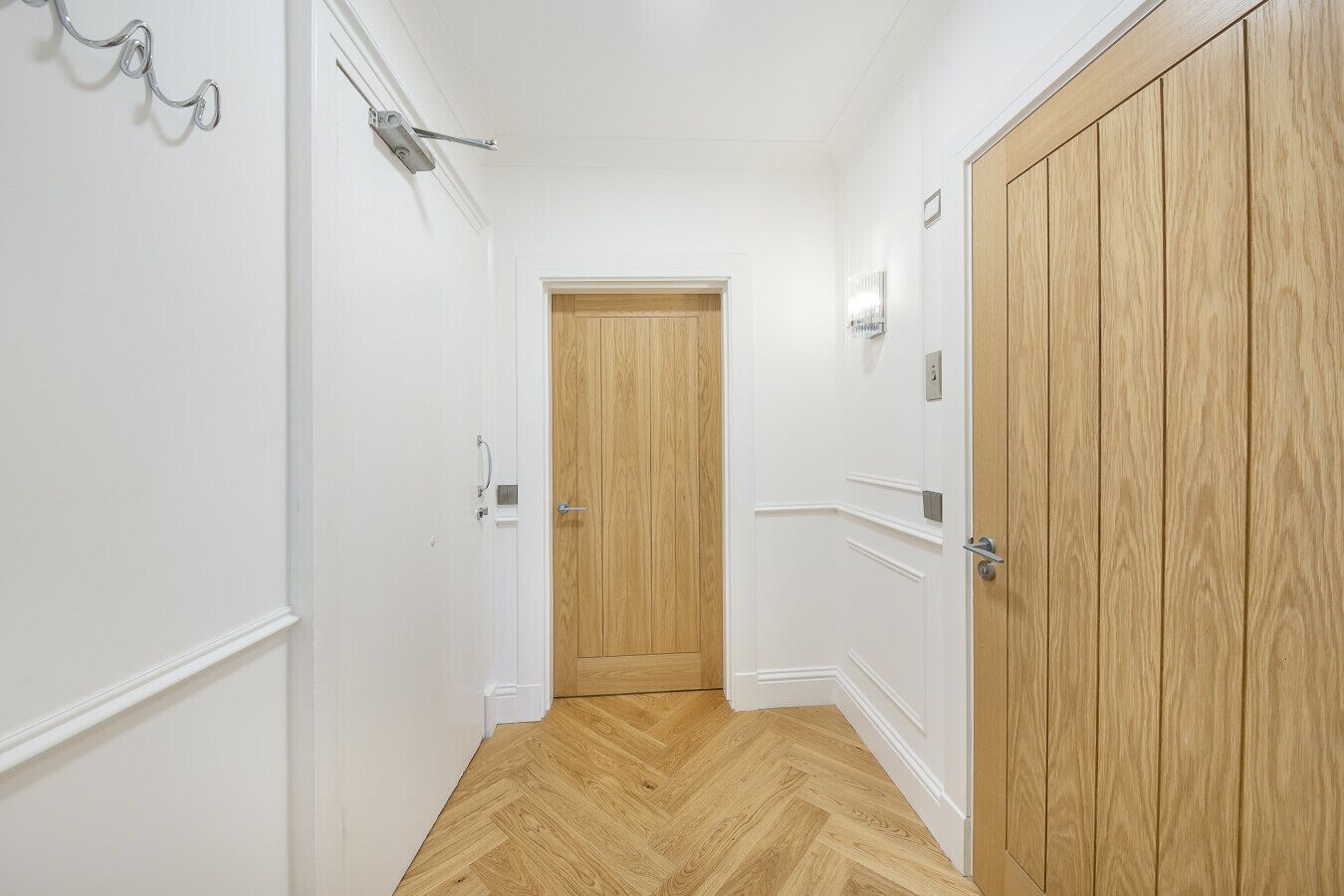 Um corredor com paredes brancas, uma porta de madeira no final e um chão com padrão herringbone.
