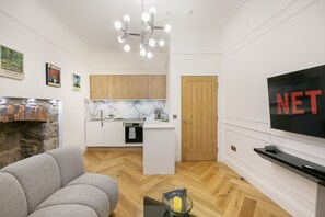A modern living room with a sofa, a TV, a fireplace, a kitchen area with wooden cabinets, and a herringbone floor pattern. - Crown Street Suite 3 - Grampian Lettings (Aberdeen)