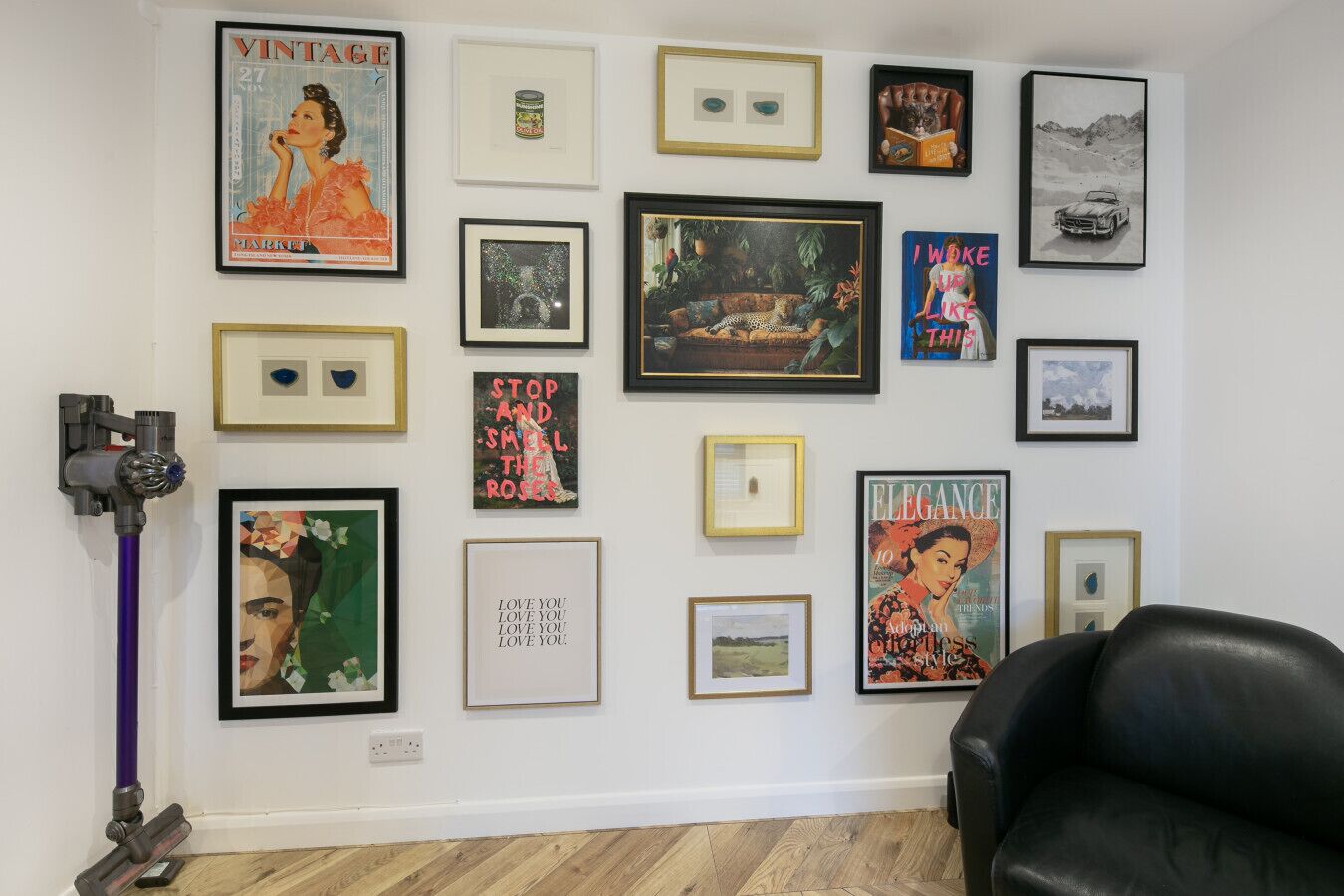 Ein Zimmer mit einer weißen Wand, die verschiedene gerahmte Kunstwerke zeigt, einem schwarzen Ledersessel und einem violetten Reinigungsgerät.