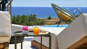 Una poltrona prendisole con vista sull'oceano, un tavolo con bevanda e un fiore, e una sedia con vista sulla piscina.