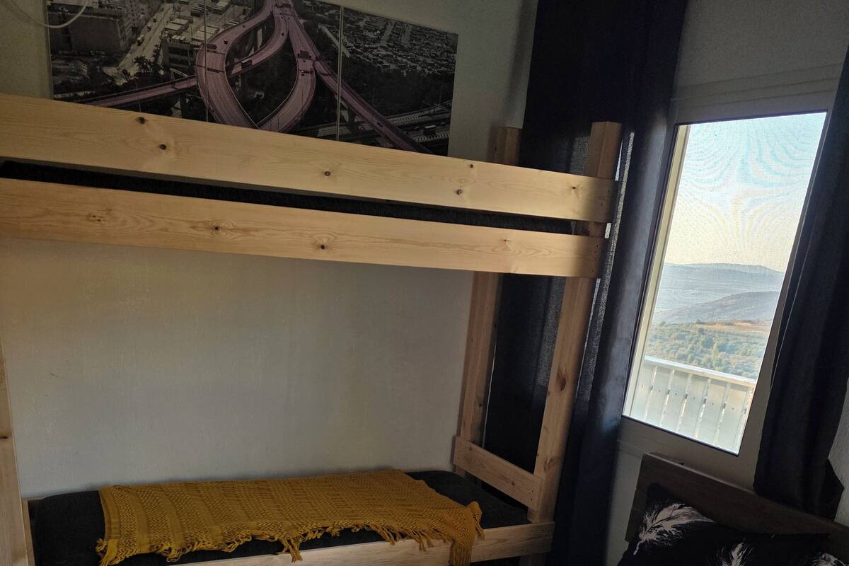 Una cama litera con una manta amarilla, una cama gris con una toalla doblada, y una ventana con vistas a un puente y montañas.