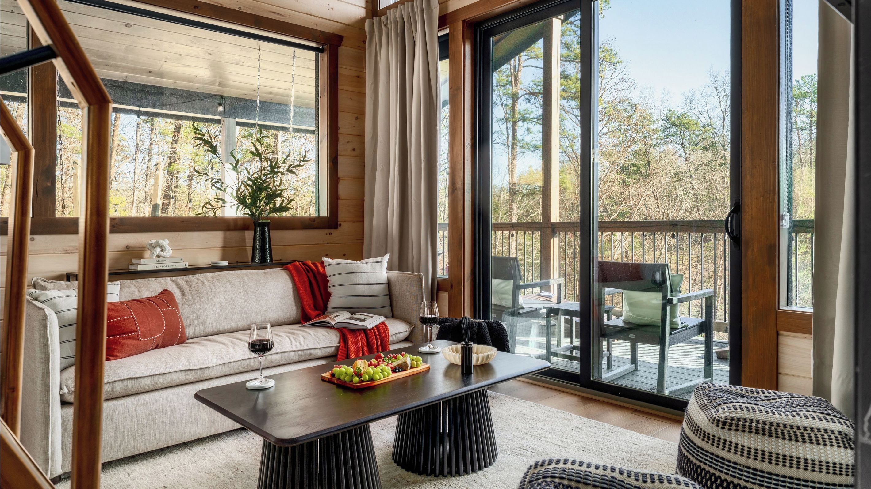 Interior acolhedor de uma cabana com sofá, mesa de jantar e uma grande janela com vista para um deck.