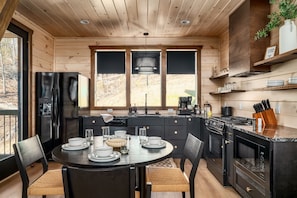 A modern kitchen with wooden cabinets, a black refrigerator, a dining table set with plates and glasses, and a large window offering a view of the outdoors. - Romantic + Luxury Cabin • Hot tub, views and more! (Sevierville)