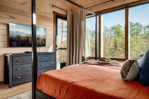 A bedroom with a large window, a bed with a red blanket, a dresser, and a painting on the wall. - Romantic + Luxury Cabin • Hot tub, views and more! (Sevierville)
