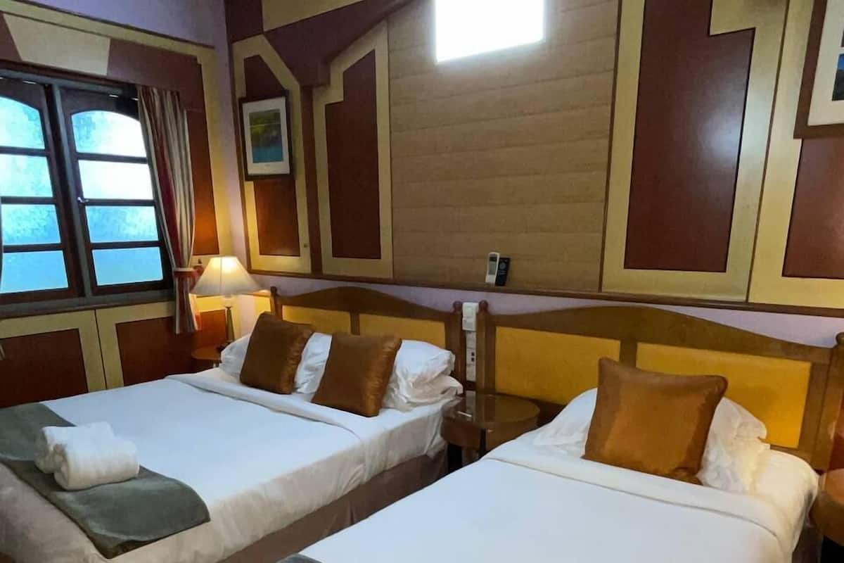 Quarto de hotel com duas camas, cada uma com lençóis brancos e almofadas marrons, uma mesa de cabeceira com abajur e uma janela com cortinas.