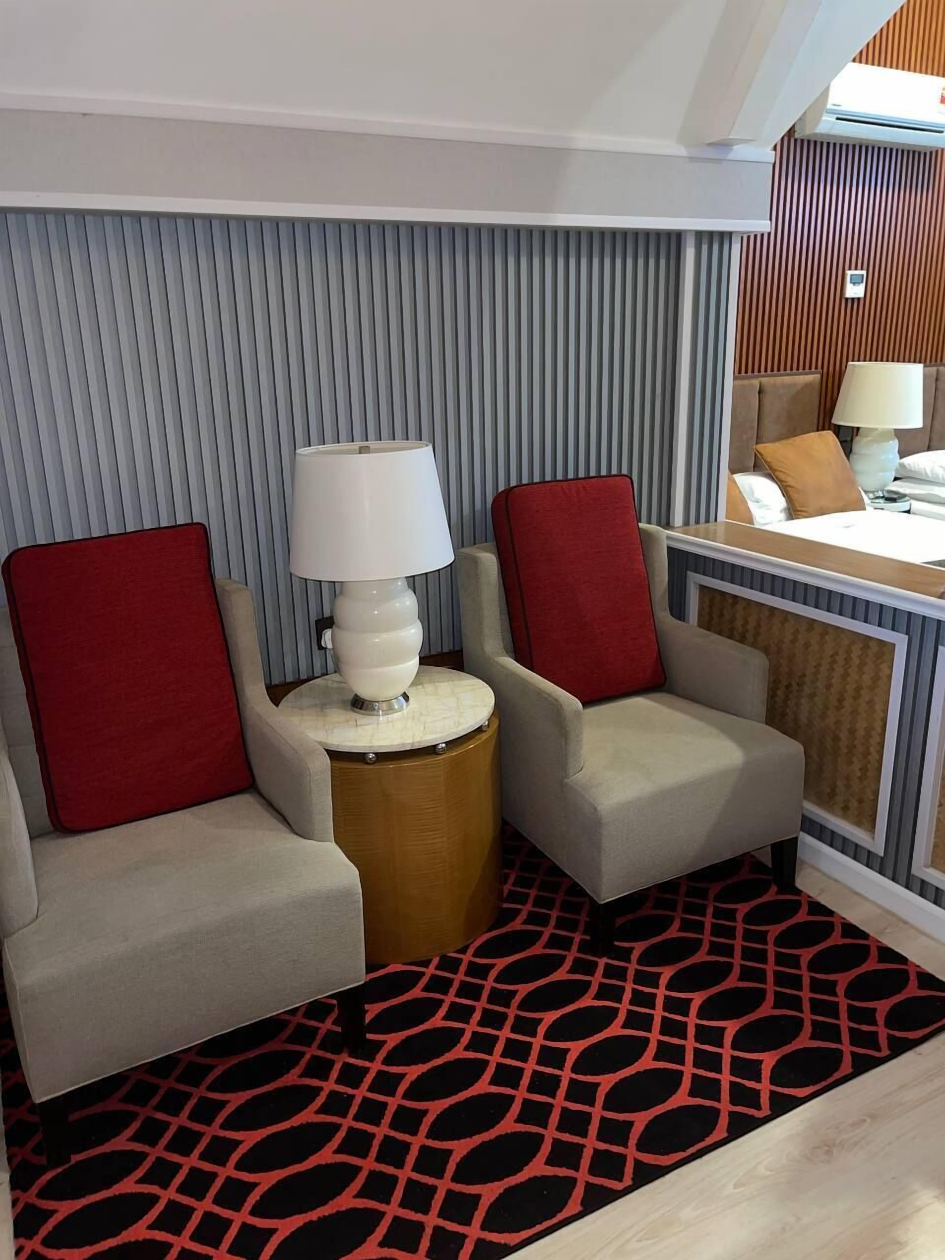 A hotel room with two armchairs, a side table with a lamp, a patterned rug, and a bed with a headboard.