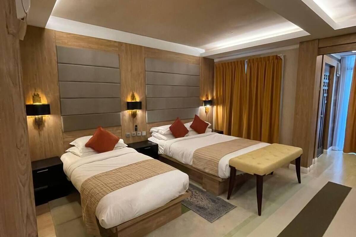 Quarto de hotel moderno com duas camas, um banco e uma janela grande com cortinas.