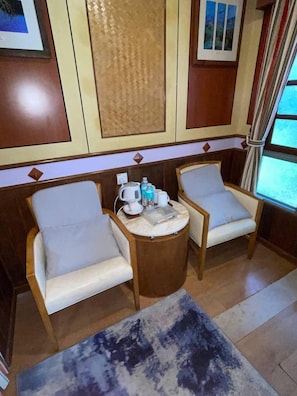 Ein Zimmer mit zwei Polstersesseln, einem kleinen runden Tisch mit Wasserkocher, Tassen und einer Wasserflasche sowie einem großen Teppich.