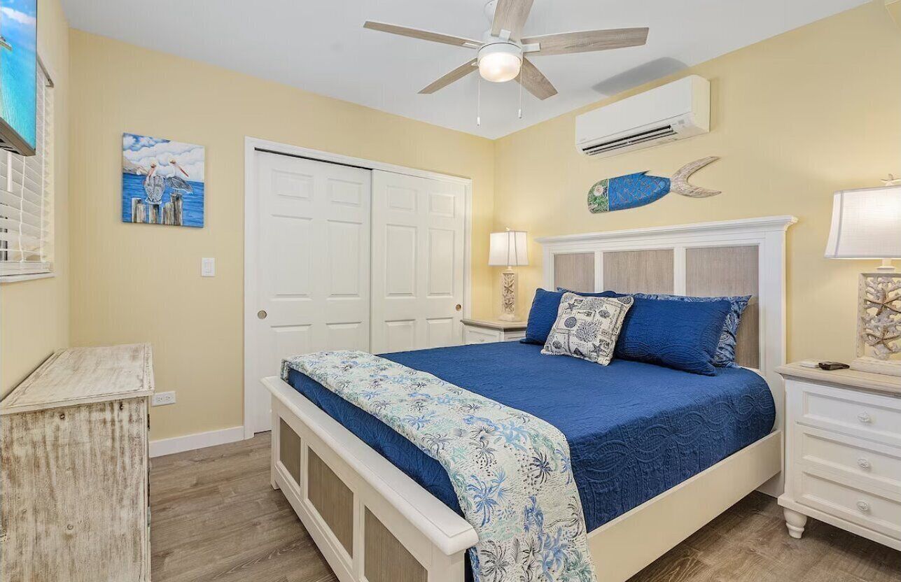 A bedroom with a bed, a ceiling fan, an air conditioning unit, a white door, and a small table with a lamp.