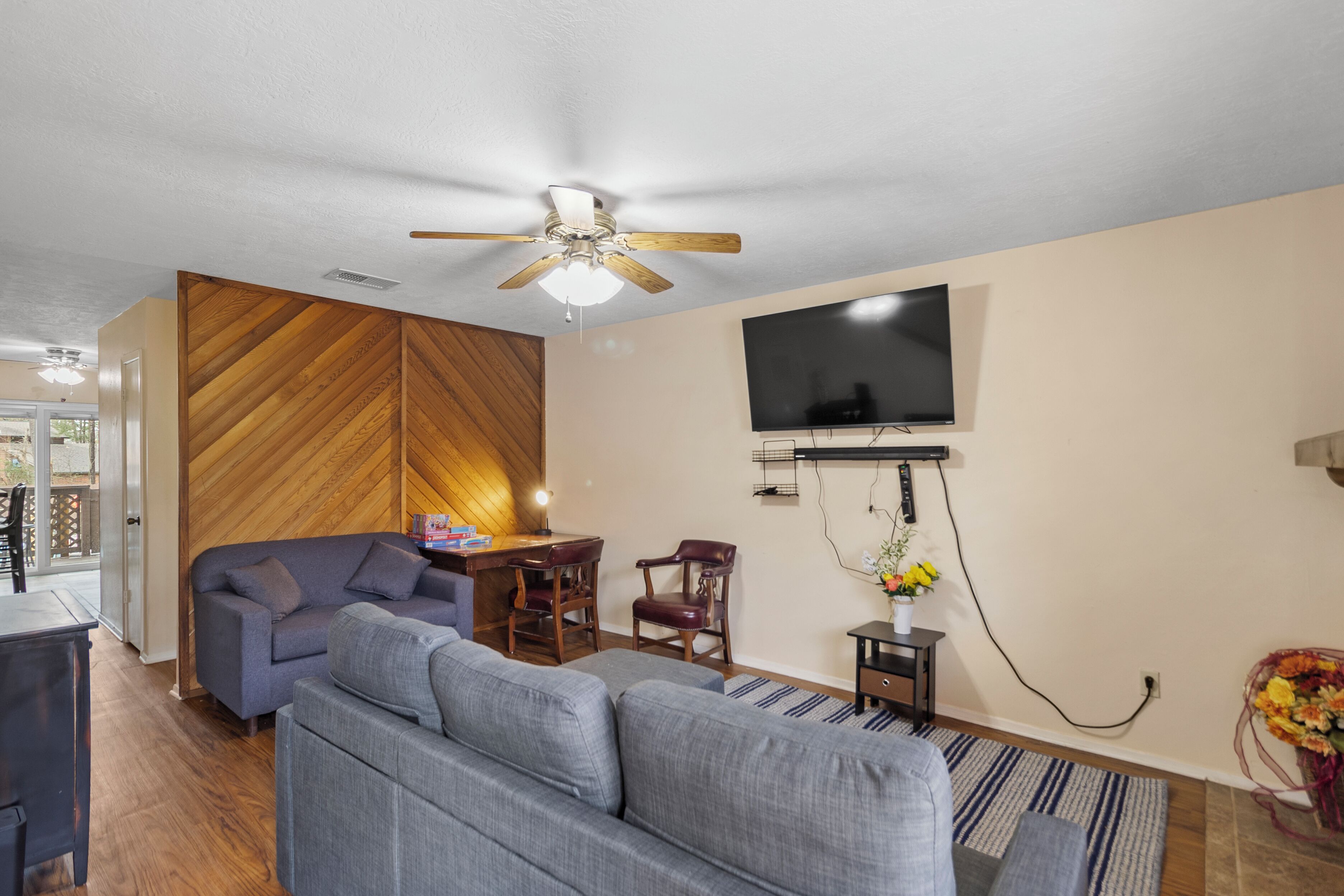 Un salon moderne avec un canapé gris, un mur d’accent en bois, un ventilateur de plafond, un téléviseur fixé au mur et une petite table avec des chaises.