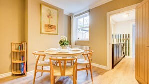 Een eetkamer met een ronde tafel voor vier personen, omringd door houten stoelen. Links is een boekenkast zichtbaar, en rechts een raam met uitzicht op buiten.