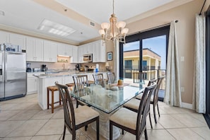 A modern kitchen with white cabinets, stainless steel appliances, a glass dining table, and a large window with a balcony view. - Phoenix III 3159: Beach Access (Orange Beach)