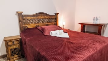 A neatly made bed with a red blanket and a rolled towel, a wooden nightstand with a drawer, a bedside lamp, and a small wooden table with two water bottles.
