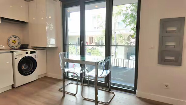 Une cuisine moderne équipée d’un lave-linge, d’une table en verre avec des chaises et d’une grande porte-fenêtre coulissante donnant sur un balcon.