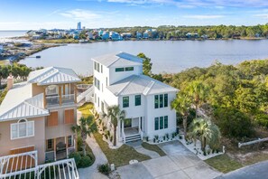 A coastal property with a large white house featuring a balcony, a smaller attached structure, and a view of the sea and a distant city skyline. - Lakefront with Pool and Gulf Views (Santa Rosa Beach)