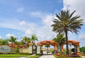 A gated entrance with a palm tree, a sign with a corn symbol, and a building with a red-tiled roof. - 6BR house near Disney with private pool (Davenport)