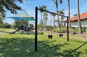 A playground with swings and a slide, surrounded by palm trees and a building with a red roof. - 6BR house near Disney with private pool (Davenport)