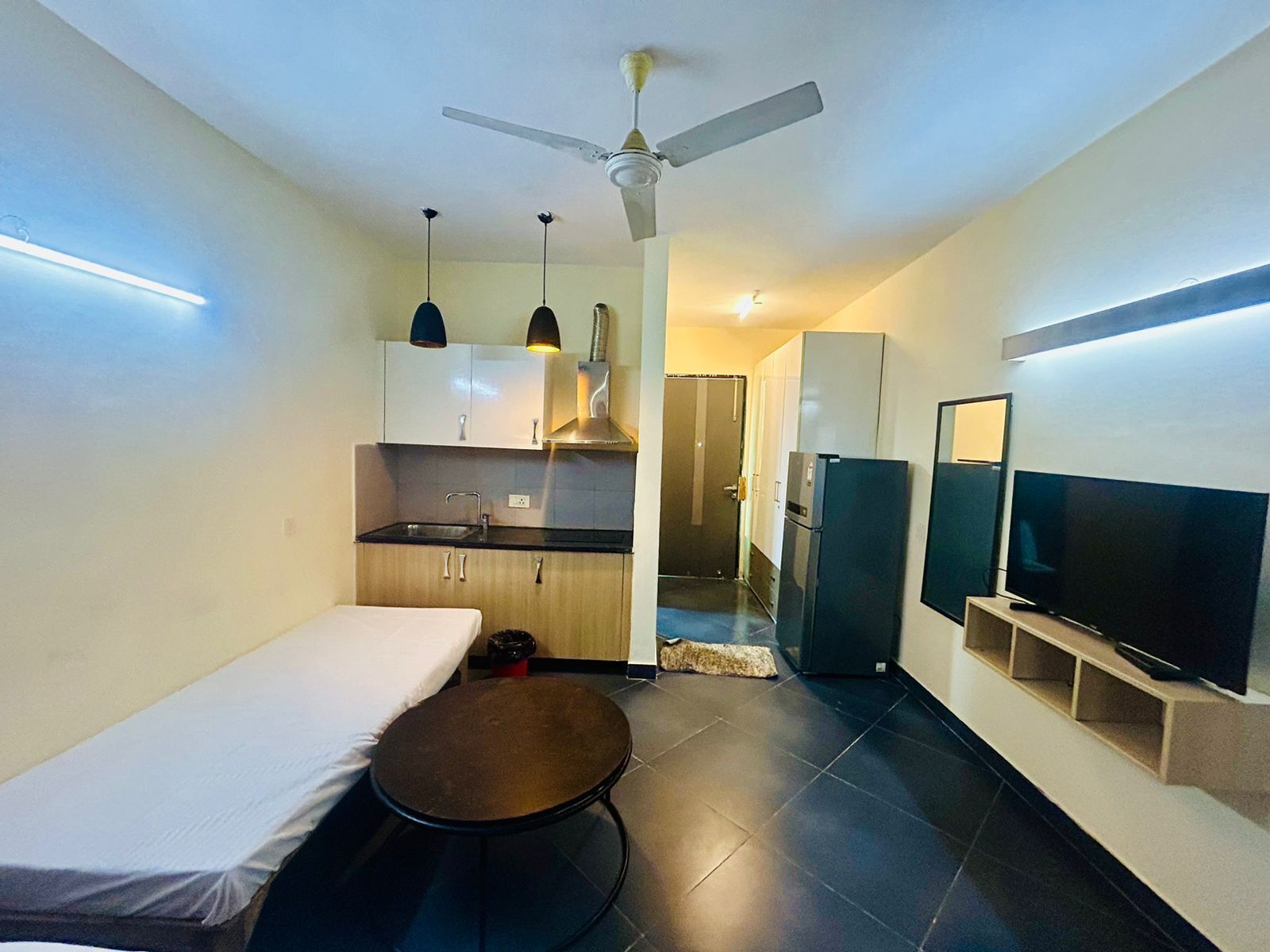 A compact hotel room with a bed, a small table, a kitchenette with a fridge, a TV, and a ceiling fan.