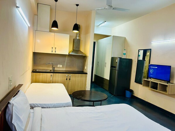 A hotel room with two beds, a kitchenette, a refrigerator, a television, and a ceiling fan. - Samriddhi Studio Apartment (New Delhi)